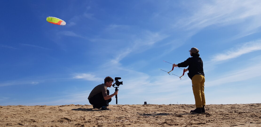 twee personen op het strand tegen een blauwe lucht. De ene is aan het vliegeren en de andere heeft een camera vast.